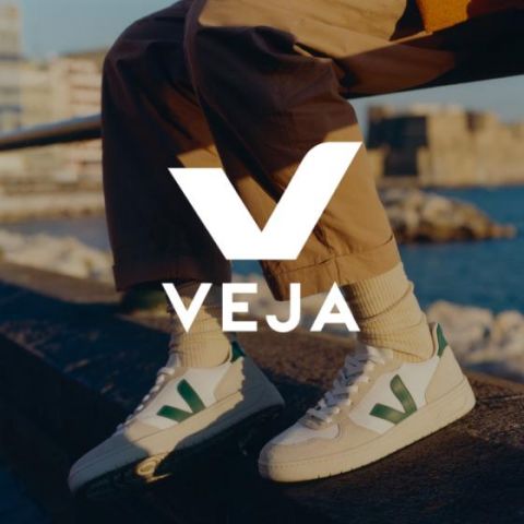 Eine Person in beiger Hose und weißen Veja-Sneakern mit grünen Akzenten sitzt auf einem Felsvorsprung am Wasser. Das Abendlicht wirft einen warmen Schein auf das Bild. Das Veja-Logo ist prominent in der Bildmitte zu sehen.