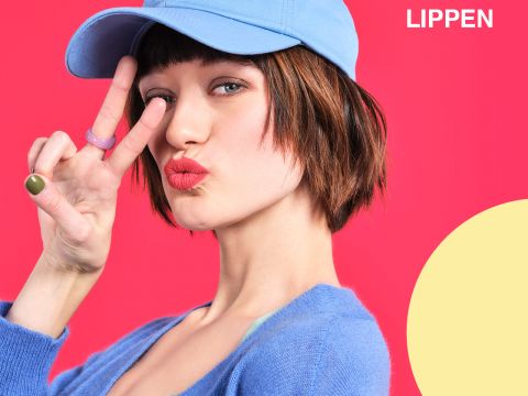 Frau mit blauer Mütze und Pullover posiert vor einem leuchtend rosa Hintergrund und macht mit einer Hand ein Peace-Zeichen. Oben rechts steht das Wort „LIPPEN“ und in der unteren rechten Ecke befindet sich ein gelber Halbkreis.
