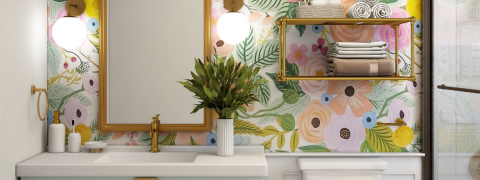 Ein lebendiges Badezimmer mit Blumentapete verfügt über ein weißes Waschbecken mit goldenem Wasserhahn, über dem ein rechteckiger Spiegel und zwei Kugellampen hängen. Auf einem goldenen Regal liegen gefaltete Handtücher und Toilettenartikel, und auf der Waschbeckenablage steht eine Vase mit üppigem Grün.