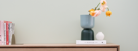 Ein minimalistischer Raum mit einem Holzregal, auf dem ein Stapel Bücher und zwei Vasen stehen; eine ist groß und blau mit rosa und gelben Blumen, die andere ist klein und weiß. Die Wand ist in einem sanften Grünton gestrichen.