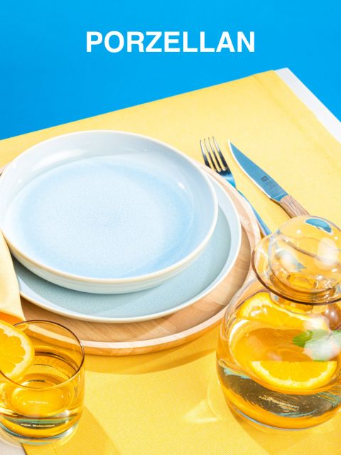 Ein heller Tisch mit zwei gestapelten blauen Tellern auf einem Holztablett, daneben Gabel und Messer. Ein Glaskrug und ein Glas, beide mit Orangenwasser gefüllt, stehen auf einer gelben Tischdecke. Darüber ist das Wort „Porzellan“ zu sehen.