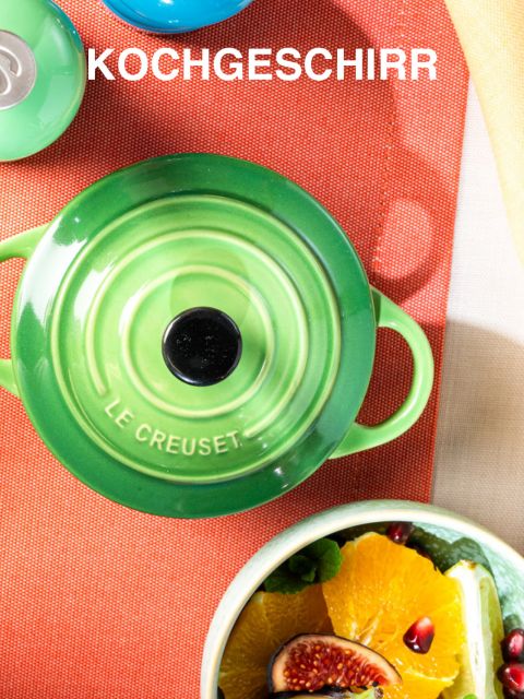 Auf einer leuchtend orangefarbenen Oberfläche steht ein grüner Le Creuset-Topf, über dem „Kochgeschirr“ steht. Daneben steht eine Schale mit Obst, darunter Orangen und Feigen, die der Szene einen Farbtupfer verleihen.