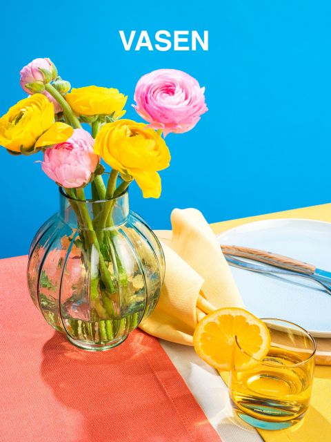 Eine durchsichtige Vase mit leuchtend gelben und rosa Blumen steht auf einer orangefarbenen Tischdecke neben einem Glas Zitronenwasser. Im Hintergrund sind eine gelbe Serviette und ein Teller mit Besteck vor einem leuchtend blauen Hintergrund zu sehen.