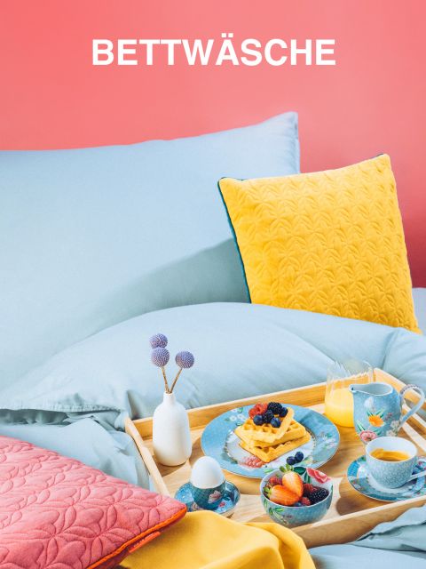 Ein gemütliches Bett mit hellblauer Bettwäsche und einem leuchtend gelben Kissen vor einer rosa Wand. Auf einem Holztablett steht ein Frühstücksset mit Waffeln, Obst, Saft und Tee auf bunten Tellern. Oben steht das Wort „Bettwäsche“.