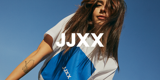 Frau mit langen Haaren und einem weiß-blauen JJXX-T-Shirt steht unter einem klaren blauen Himmel. Sie hat ein Tattoo auf dem Arm.