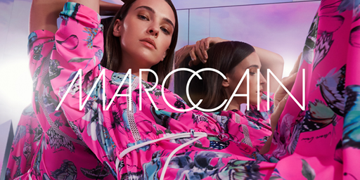 Eine Frau in einem leuchtend rosa-blau gemusterten Outfit posiert selbstbewusst. Der Markenname „Marc Cain“ ist prominent über dem Bild zu sehen.