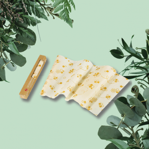 Auf hellgrünem Untergrund ist ein Bienenwachstuch mit gelbem Blumenmuster ausgefaltet abgebildet. Daneben steht eine passende Schachtel. Das Tuch ist von sattgrünen Eukalyptusblättern umgeben.