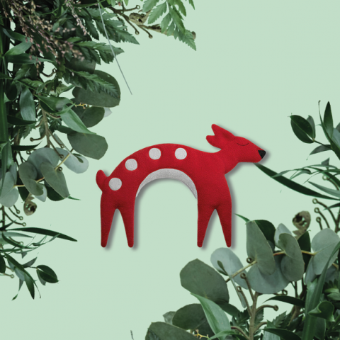 Ein rotes, stilisiertes Spielzeughirsch mit weißen Flecken steht vor einem hellgrünen Hintergrund. Grüne Blätter rahmen die Seiten des Bildes ein.