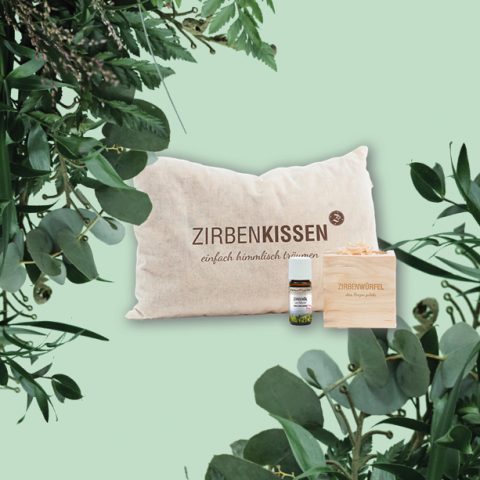 Auf einer Holzkiste mit der Aufschrift „Zirbenwürfel“ liegt ein Kissen mit der Aufschrift „Zirbenkissen“, dazu eine kleine Flasche. Der Hintergrund besteht aus grünem Blattwerk und einem hellgrünen Hintergrund. Der Text unter „Zirbenkissen“ lautet „einfach himmlisch träumen“.