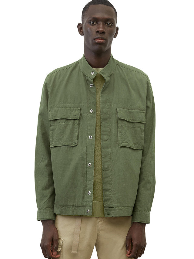 Eine Person trägt eine grüne Jacke mit zwei Brusttaschen und einem grünen Pullover. Sie trägt außerdem beige Hosen und steht vor einem weißen Hintergrund.