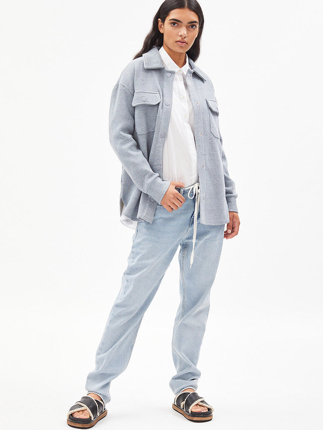 Eine Person steht selbstbewusst da und trägt eine hellgraue, übergroße Jacke über einem weißen Hemd, dazu lockere hellblaue Jeans und schwarze Sandalen. Der Hintergrund ist schlicht weiß.
