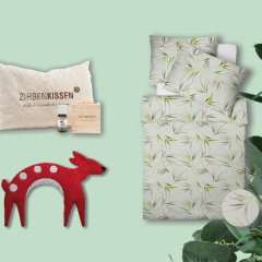 Ein Display mit einem beigen Kissen mit der Aufschrift „Zirbenkissen“ mit kleiner Flasche und Tasche, einer roten Deko-Hirschfigur mit weißen Punkten und einem Bettset mit Blättermuster. Rechts ist eine grüne Blattpflanze teilweise zu sehen.