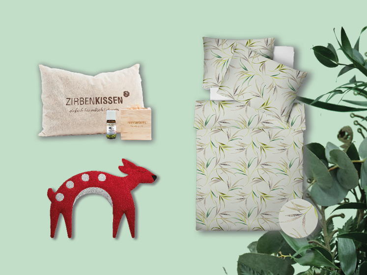Ein Display mit einem beigen Kissen mit der Aufschrift „Zirbenkissen“ mit kleiner Flasche und Tasche, einer roten Deko-Hirschfigur mit weißen Punkten und einem Bettset mit Blättermuster. Rechts ist eine grüne Blattpflanze teilweise zu sehen.