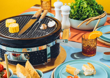 Ein farbenfroher Picknicktisch mit einem tragbaren Grill, auf dem Mais und Knoblauch gegrillt werden. Um den Grill herum stehen Teller mit geschnittenem Brot, frischem Salat und Gläsern mit Eistee und Orangenscheiben. Bunte Teller und eine fröhliche, sonnige Atmosphäre.