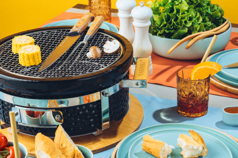 Ein farbenfroher Picknicktisch mit einem tragbaren Grill, auf dem Mais und Knoblauch gegrillt werden. Um den Grill herum stehen Teller mit geschnittenem Brot, frischem Salat und Gläsern mit Eistee und Orangenscheiben. Bunte Teller und eine fröhliche, sonnige Atmosphäre.
