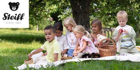 Eine Gruppe Kinder sitzt auf einer Decke im Freien, umgeben von Bäumen. Sie spielen mit Körben und Stofftieren. Das Steiff-Logo befindet sich in der oberen linken Ecke.