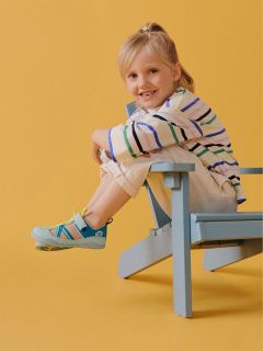 Lächelndes Kind in gestreiftem Hemd und heller Hose sitzt seitwärts auf einem blauen Holzstuhl vor gelbem Hintergrund.