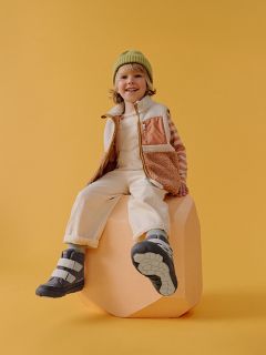 Ein lächelndes Kind mit grüner Mütze, gestreiftem Hemd und Weste sitzt auf einem pfirsichfarbenen Block vor einem gelben Hintergrund und trägt Stiefel und eine helle Hose.