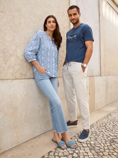 Eine Frau in einer blau gemusterten Bluse und hellen Jeans steht neben einem Mann in einem blauen T-Shirt und beigen Hosen. Sie sind draußen, lehnen an einer strukturierten Wand auf einem Steinweg und sehen beide entspannt und lässig aus.
