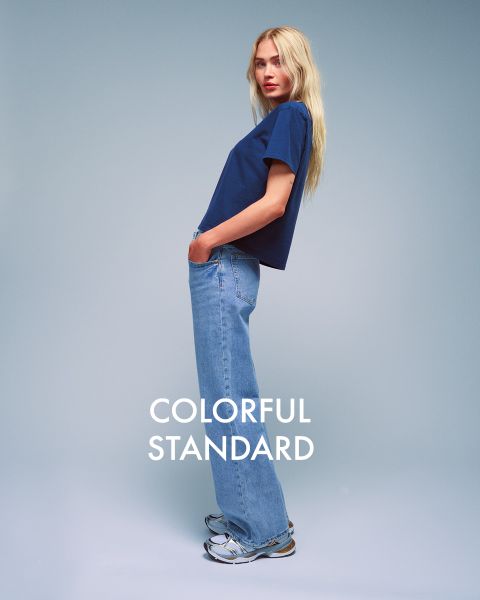 Eine Person mit langen blonden Haaren steht seitlich vor einem blauen Hintergrund. Sie trägt ein dunkelblaues T-Shirt, hellblaue Jeans und Turnschuhe. Der Text „COLORFUL STANDARD“ ist in Weiß dargestellt.