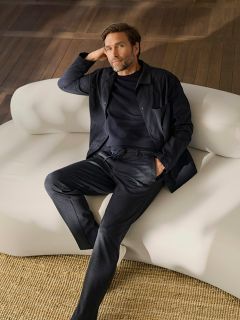 Ein bärtiger Mann in dunkler Kleidung sitzt entspannt auf einem weißen, geschwungenen Sofa, das auf einem gewebten Teppich in einem Raum mit Holzboden steht.
