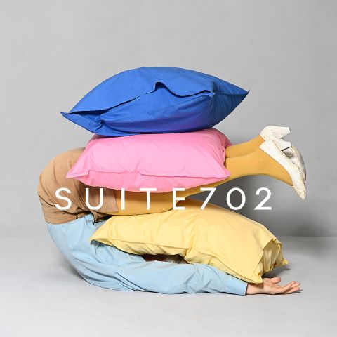 Eine Person liegt unter drei großen Kissen - blau, rosa und gelb -, die Beine ragen heraus, sie trägt gelbe Strumpfhosen und weiße Schuhe. Der Text lautet "SUITE 702.