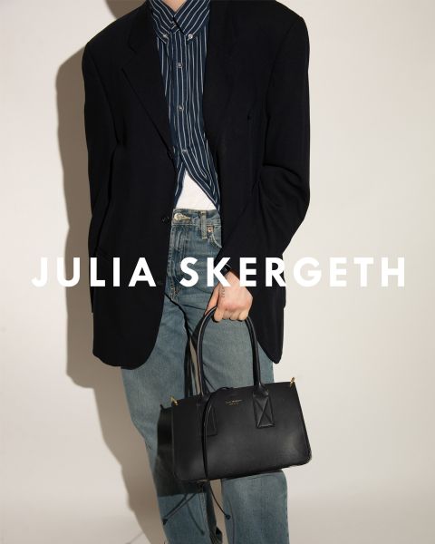 Person in einem schwarzen Blazer über einem gestreiften Hemd und blauen Jeans, die eine schwarze Handtasche hält. Der Text „JULIA SKERGETH“ ist deutlich sichtbar über das Bild gelegt.
