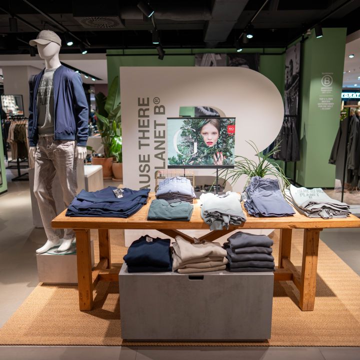 Auslage eines Bekleidungsgeschäfts mit gefalteten Hemden und Hosen auf einem Holztisch, einer Schaufensterpuppe in Freizeitkleidung und einem Schild "Use There Is No Planet B".