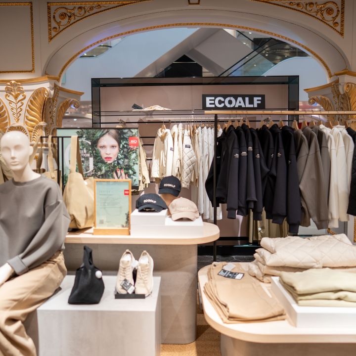 Ein Bekleidungsgeschäft mit einer Schaufensterpuppe in neutraler Kleidung, gefalteten Kleidern, Hüten, Taschen und einem Jackenständer unter einem "ECOALF"-Schild.