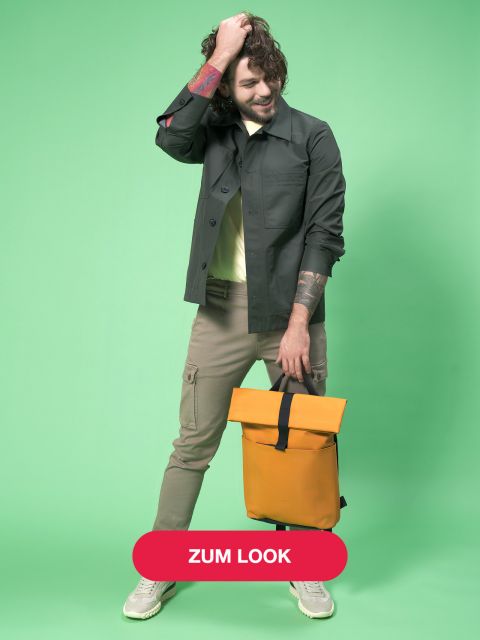Mann mit lockigem Haar, dunkler Jacke und beiger Hose hält einen orangefarbenen Rucksack vor grünem Hintergrund. Er hat Tattoos auf den Armen und trägt grüne Turnschuhe. Auf einem roten Button an der Unterseite steht „Zum Look“.