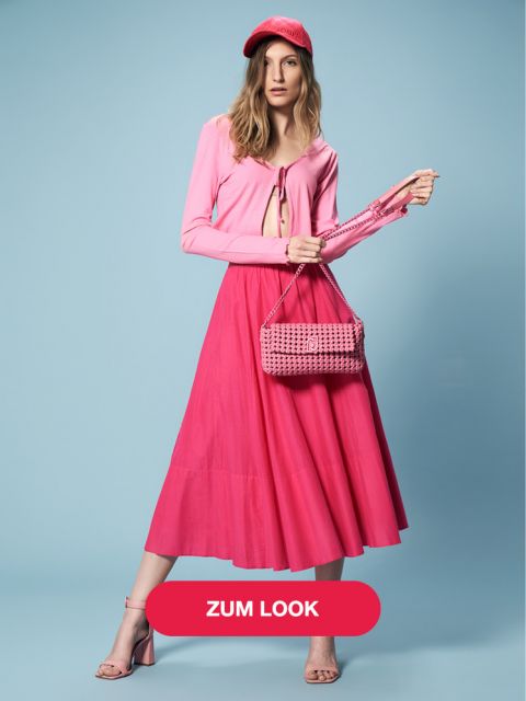 Eine Frau in einem rosa Outfit posiert vor einem blauen Hintergrund. Sie trägt ein langärmliges Oberteil, einen Midirock, eine Mütze und Sandalen, alles in Rosa. Sie hält eine gewebte Tasche und auf einem Knopf an der Unterseite ist der Text „ZUM LOOK“ zu sehen.