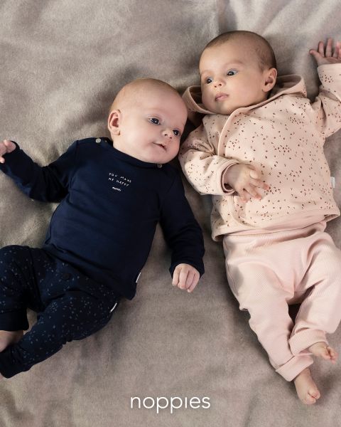 Zwei Babys liegen auf einer weichen Decke. Das Baby links trägt ein marineblaues Outfit mit weißem Muster, während das Baby rechts ein pfirsichfarbenes Outfit mit dunkleren Sprenkeln trägt. Unten im Bild erscheint das Wort „Noppies“.