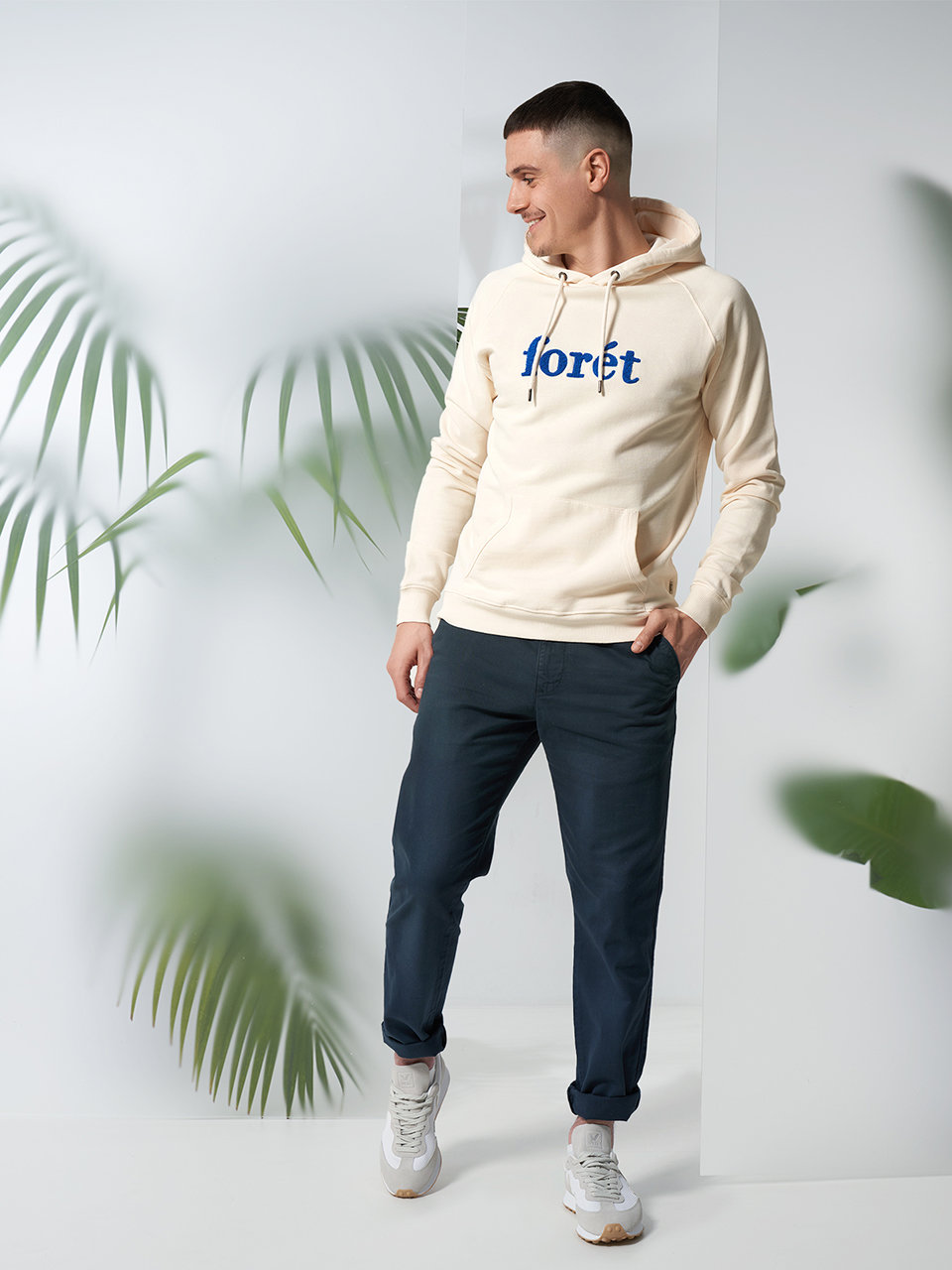Ein Mann mit einem beigen Kapuzenpullover mit der Aufschrift „Loret“, marineblauen Hosen und weißen Turnschuhen steht in einem hellen Raum mit Palmblättern im Hintergrund. Er lächelt und schaut dabei leicht zur Seite.