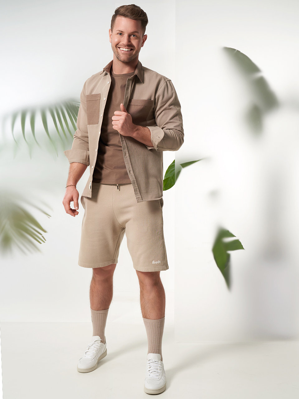 Ein Mann steht lächelnd vor einem hellen Hintergrund mit grünen Blättern. Er trägt eine beige Jacke über einem braunen Hemd, beige Shorts, lange beige Socken und weiße Turnschuhe. Seine rechte Hand steckt in der Tasche und seine linke Hand richtet seine Jacke.