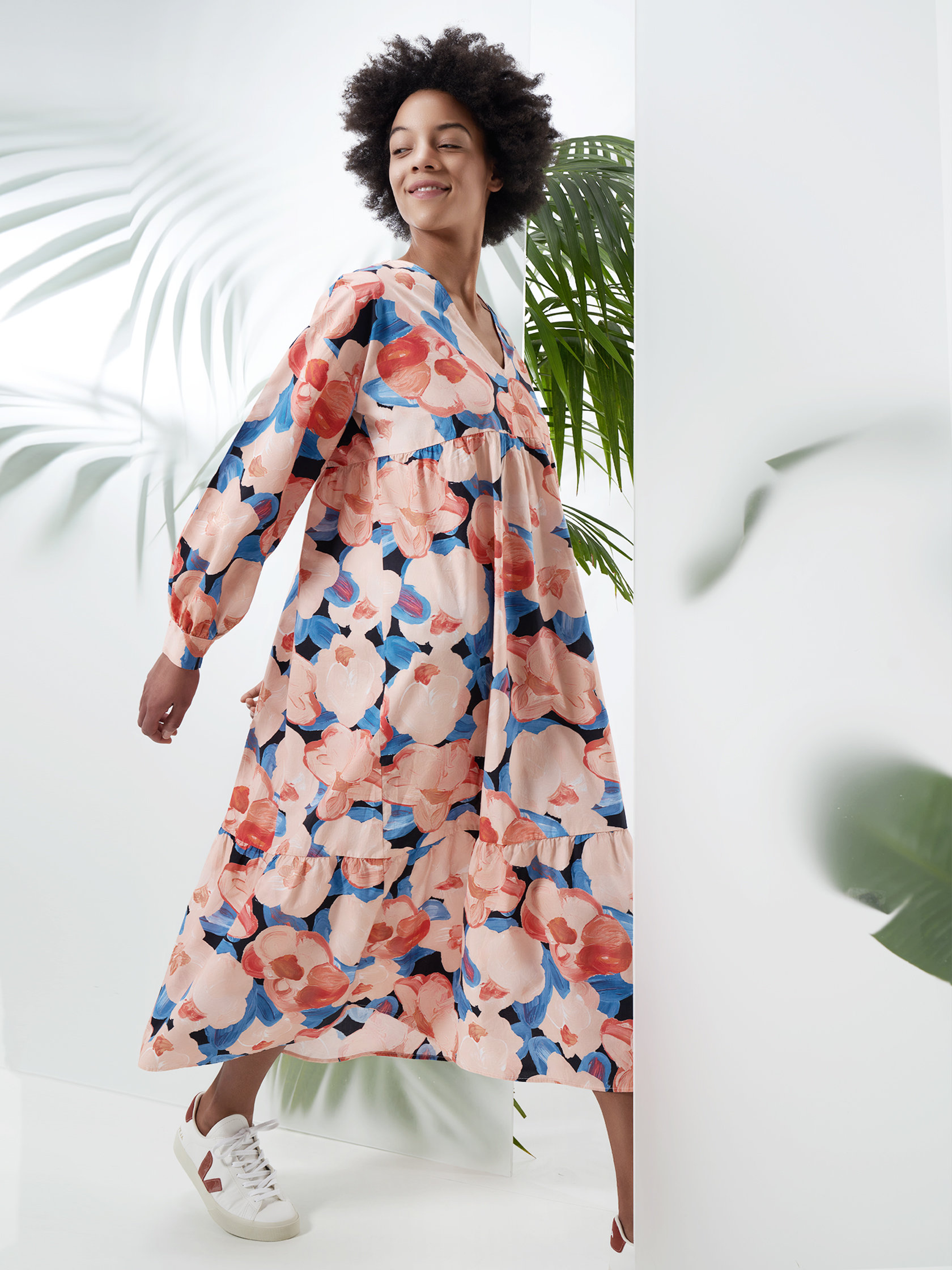 Eine Person lächelt, während sie ein langärmliges Kleid mit Blumenmuster trägt und neben großen grünen Blättern läuft. Das Kleid ist in den Farben Rosa, Blau und Weiß gehalten und sorgt so für einen lebendigen und sommerlichen Look. Sie trägt weiße Turnschuhe.