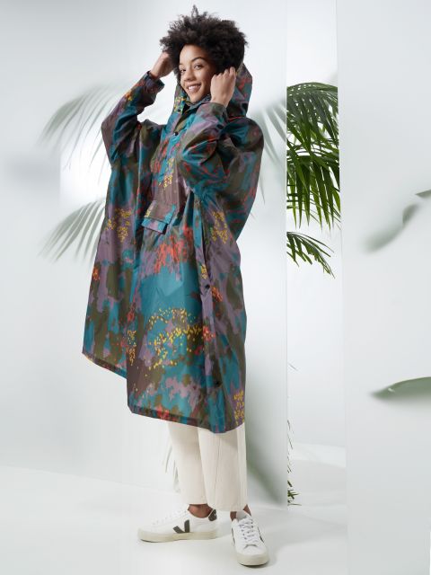 Eine Person lächelt und trägt einen farbenfrohen Regenmantel mit Blumenmuster und hochgezogener Kapuze. Sie steht in einem Haus neben grünen Blattpflanzen. Die Person hat kurzes lockiges Haar, helle Hosen und weiße Turnschuhe. Der Hintergrund ist hell und minimalistisch.