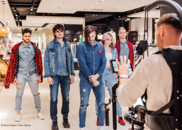 Eine Gruppe von fünf Personen in Jeans-Outfits posiert gemeinsam in einem Geschäft. Ein Fotograf, ausgerüstet mit Kamera und Ausrüstung, macht ein Foto von ihnen. Die Umgebung ist hell und modern, im Hintergrund sind verschiedene Artikel ausgestellt.