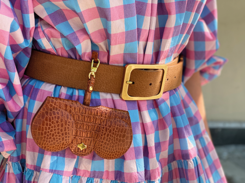 Eine Nahaufnahme einer Person, die ein kariertes Kleid in Pink, Blau und Weiß mit einem breiten braunen Ledergürtel trägt. Am Gürtel ist ein kleiner, strukturierter brauner Lederbeutel befestigt. Der Gürtel hat eine große Messingschnalle.