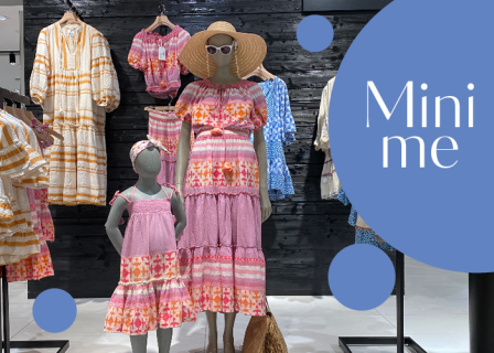 Eine Kleiderschau mit einer Schaufensterpuppe in einem bunten, gemusterten Kleid und Hut, mit einem passenden Kinderkleid auf einer anderen Schaufensterpuppe. Im Hintergrund sind weitere Kleider zu sehen. In einer Sprechblase steht „Mini me“ und blaue Kreise schmücken die Szene.