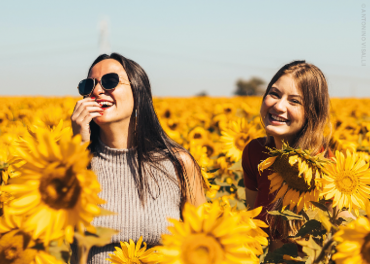 Zwei Frauen stehen lächelnd in einem leuchtenden Sonnenblumenfeld unter einem klaren blauen Himmel. Eine trägt eine Sonnenbrille und ein ärmelloses Oberteil, die andere ein kurzärmeliges Hemd. Sie scheinen einen sonnigen Tag zu genießen.