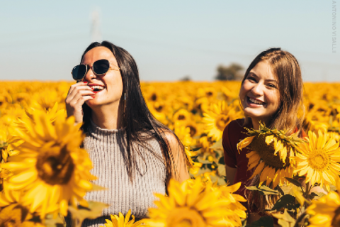 Zwei Frauen stehen lächelnd in einem leuchtenden Sonnenblumenfeld unter einem klaren blauen Himmel. Eine trägt eine Sonnenbrille und ein ärmelloses Oberteil, die andere ein kurzärmeliges Hemd. Sie scheinen einen sonnigen Tag zu genießen.