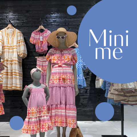 In einem Geschäft stehen Schaufensterpuppen in passenden rosa und orangefarbenen Outfits, darunter Kleider und Hüte. Im Hintergrund sind weitere Kleidungsstücke auf Kleiderbügeln zu sehen. Auf der rechten Seite ist ein „Mini me“-Schild zu sehen.