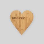 Ein herzförmiges Holzschneidebrett mit deutschem Text: „Der Mensch lebt nicht vom Brot allein. Es darf auch etwas Liebe sein.“ vor einem schlichten weißen Hintergrund.