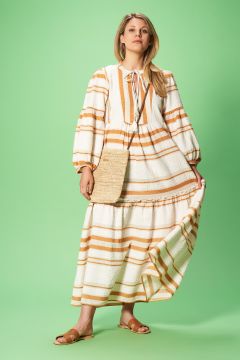 Eine Person in einem langen, weißen Kleid mit senfgelben Streifen posiert vor einem grünen Hintergrund. Sie hält einen Teil des Kleides und hat eine gewebte Umhängetasche. Sie trägt Sandalen und hat lange Haare.