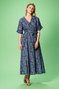 Eine Frau in einem blau gemusterten Wickelkleid steht vor einem grünen Hintergrund. Sie trägt ein geblümtes Stirnband und goldene Sandalen, ihre Hände sind locker vor der Brust verschränkt.