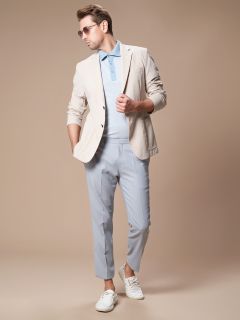 Eine Person in einem hellen Blazer, einem blauen Poloshirt, hellblauen Hosen und weißen Turnschuhen posiert vor einem beigen Hintergrund. Sie trägt eine Sonnenbrille und hat die Hände in den Blazertaschen, was eine entspannte und stilvolle Ausstrahlung ausstrahlt.