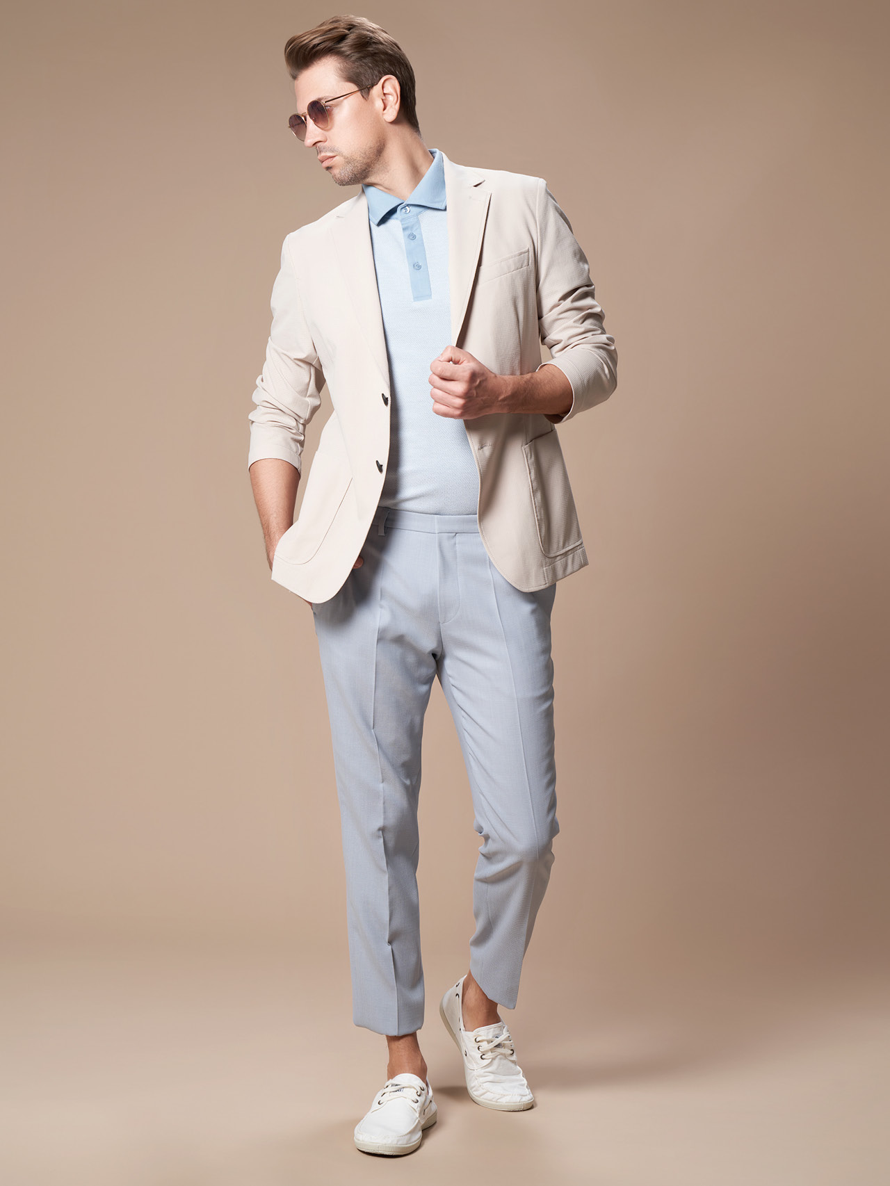 Eine Person in einem hellen Blazer, einem blauen Poloshirt, hellblauen Hosen und weißen Turnschuhen posiert vor einem beigen Hintergrund. Sie trägt eine Sonnenbrille und hat die Hände in den Blazertaschen, was eine entspannte und stilvolle Ausstrahlung ausstrahlt.