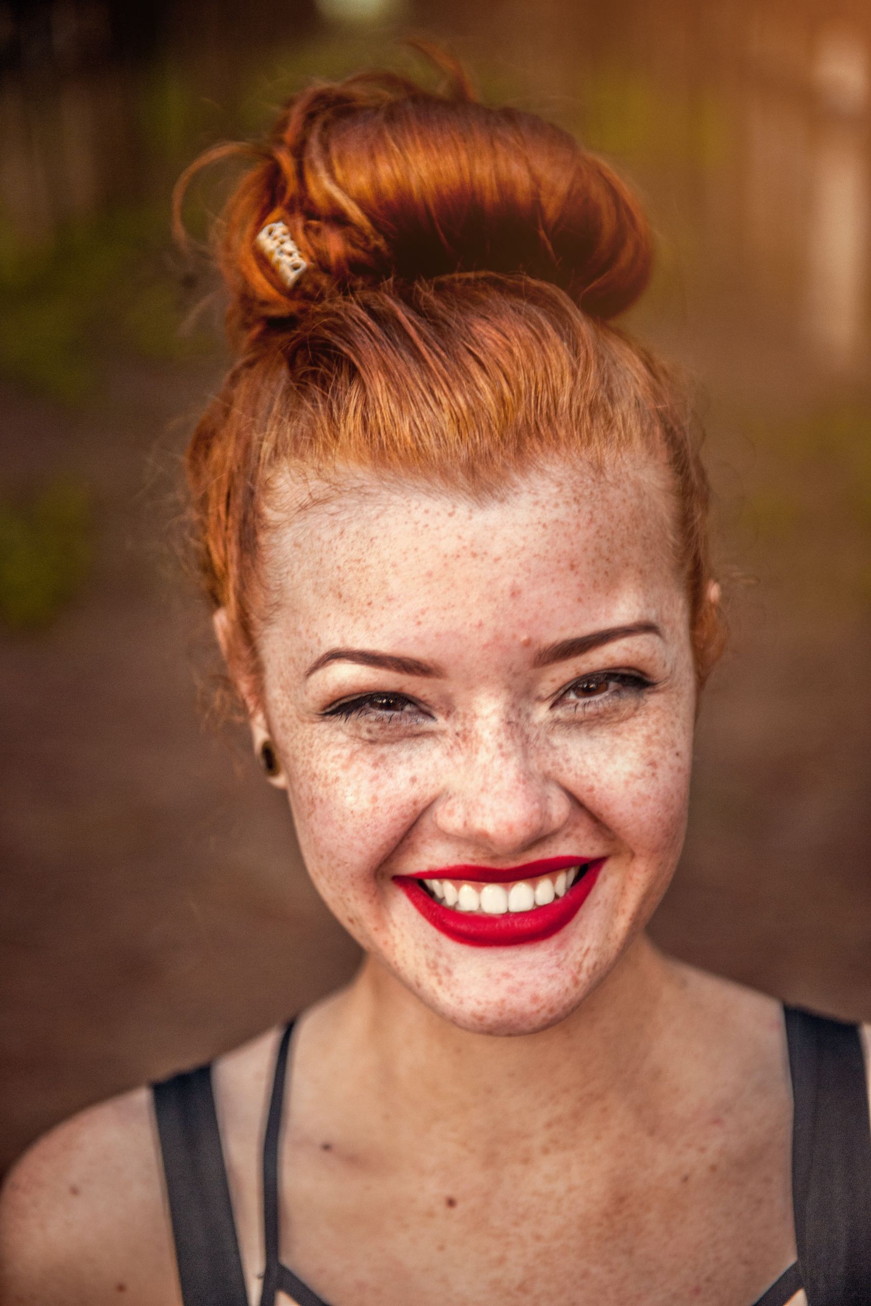 Eine Frau mit rotem Haar im Dutt lächelt strahlend und zeigt ihre Sommersprossen und ihren roten Lippenstift. Sie ist draußen, mit einem verschwommenen Hintergrund aus Grün und Sonnenlicht.