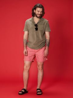 Eine Person mit langen Haaren und Tätowierungen steht vor einem roten Hintergrund, trägt ein grünes Hemd, rot-weiß gemusterte Shorts, schwarze Sandalen und hält eine Sonnenbrille.