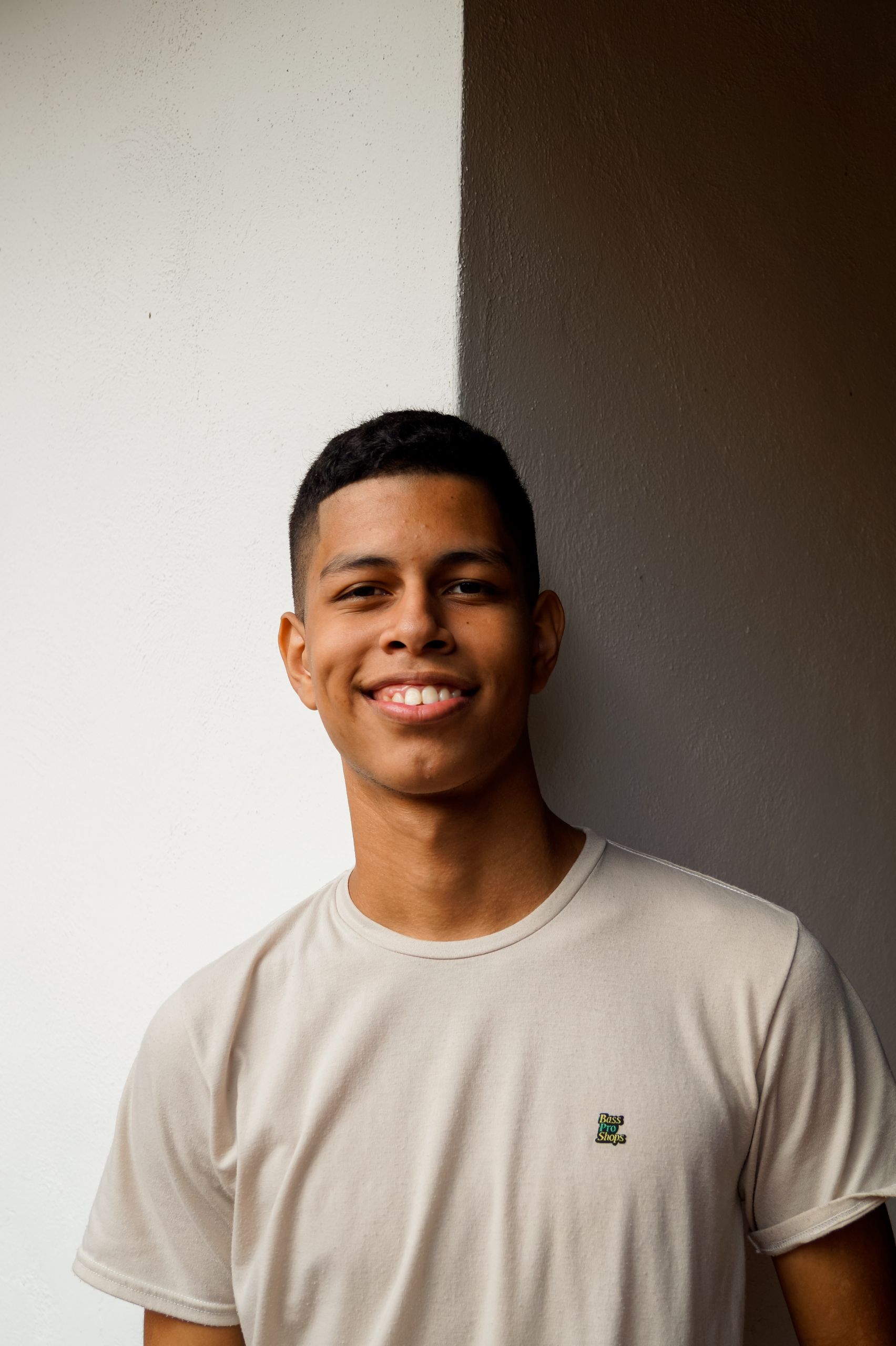 Eine junge Person mit kurzen dunklen Haaren steht lächelt vor einer weiß-grauen Wand und trägt ein hellbeiges T-Shirt.